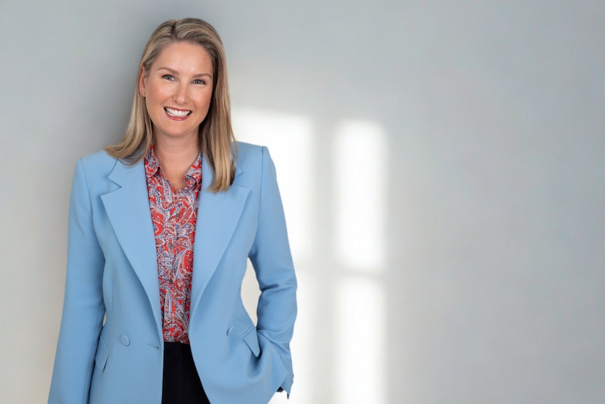 Victoria Momsen, a white woman with shoulder length blonde hair, wears a pale blue blazer and leans against a white wall
